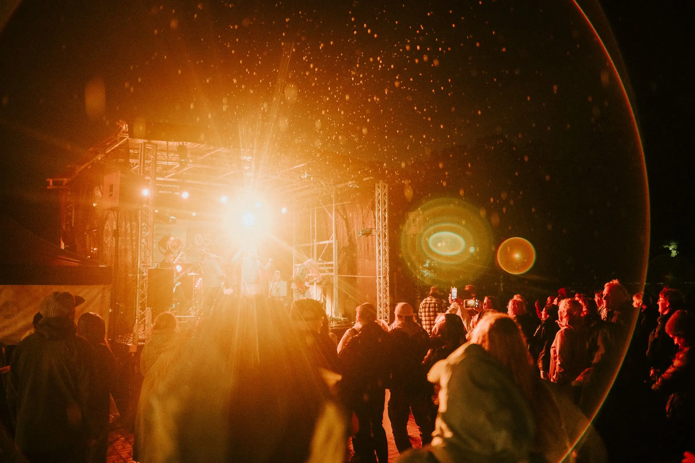 Vakara tirdziņš parkā, cilvēki pie izgaismotiem stendiem zem lieliem kokiem